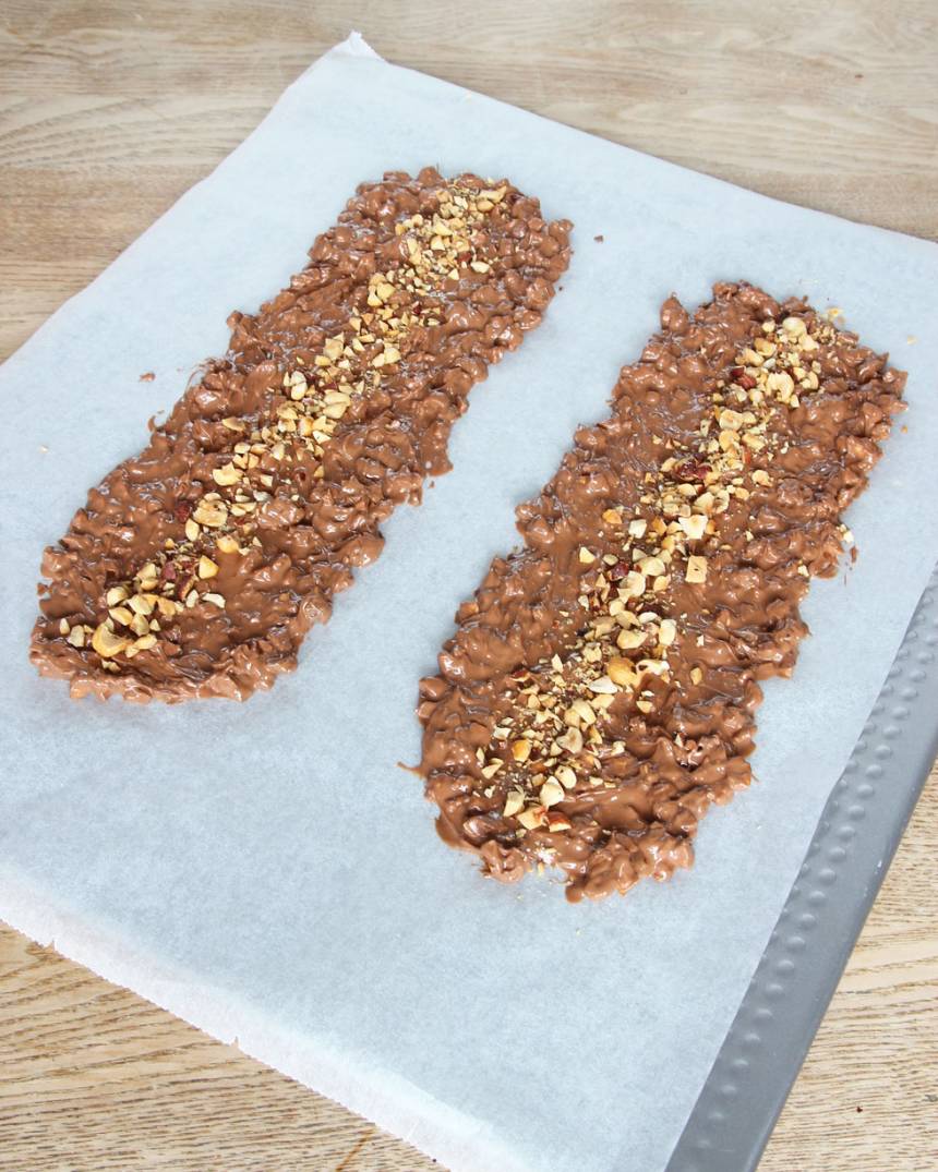 6. Bred ut chokladen till två längder på ett bakplåtspapper. Strö över resten av hasselnötterna. Låt chokladen stelna i kylen. Förvara bräcken i kylen för den kan mjukna i rumsvärme.